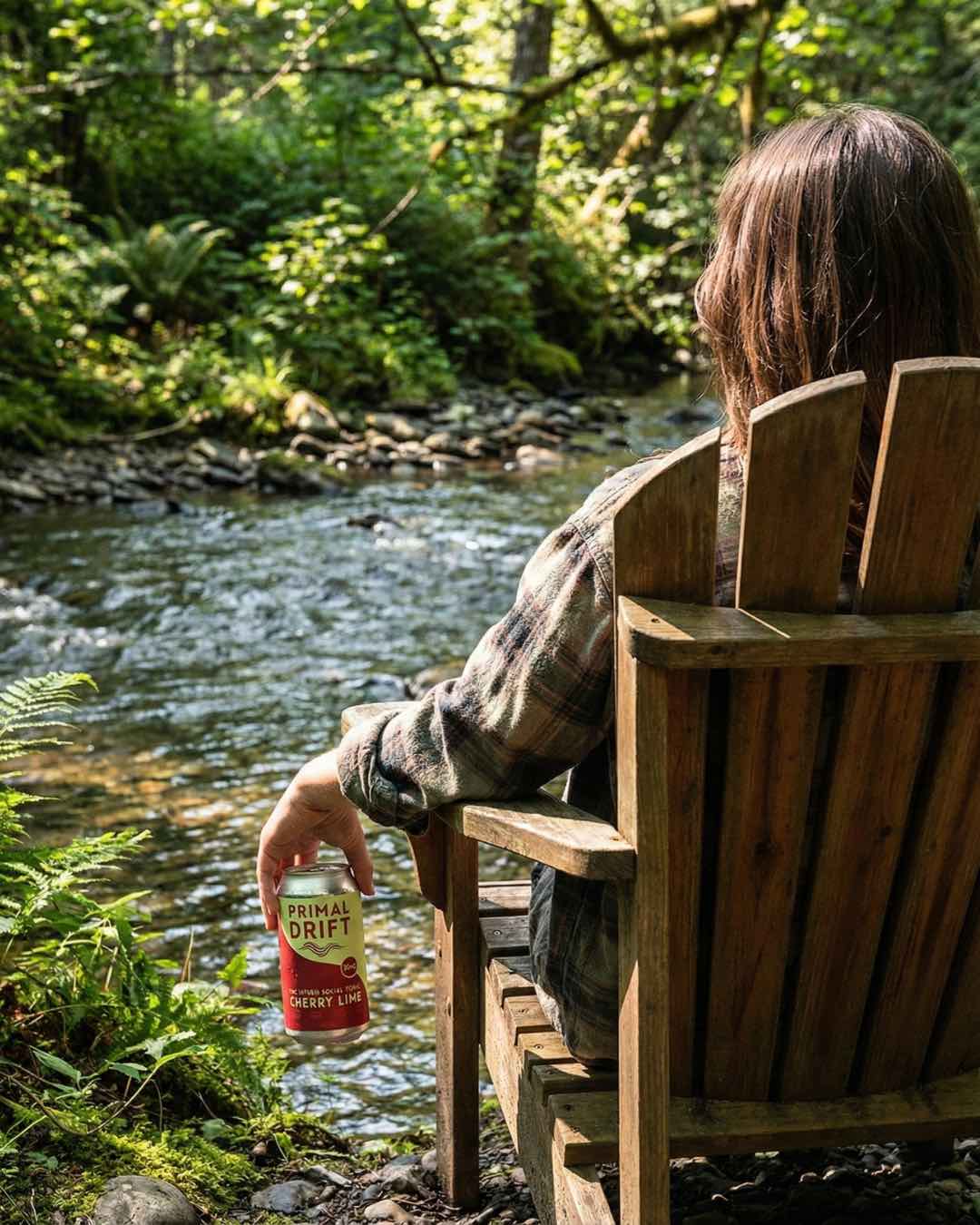 Person sitting in a chair by a river holding a Primal Drift Cherry Lime social tonic can